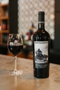 A bottle of red wine with a label depicting a biker riding a vintage motorcycle stands next to a full glass of red wine