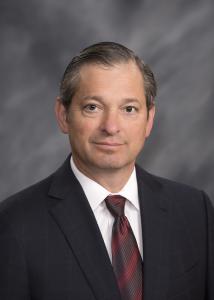 Headshot of Dr. Greg Vigna in a suit with a maroon tie in front of a grey background