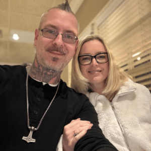A smiling couple, with the man displaying tattoos and a necklace and the woman wearing glasses and a white fleece, pose together indoors.