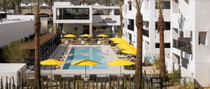 A modern hotel courtyard features a rectangular swimming pool, yellow umbrellas, lounge chairs, and palm trees, surrounded by a cactus fence and flanked by white buildings.